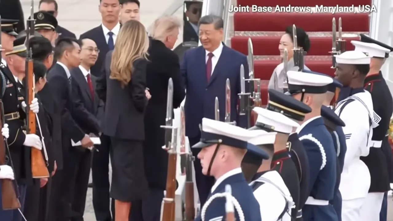 Trump and Xi stand near an aircraft stairway while officials greet them and uniformed personnel stand in formation